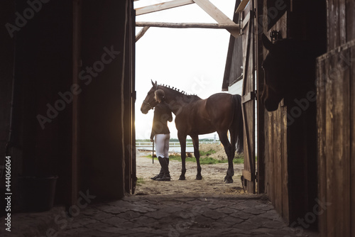 Back view of a Horse woman standing with her dark Bay horse outside the stable. Posing for the camera. View from the door of the stable. Another horse can be seen looking out from its stall. Daytime.