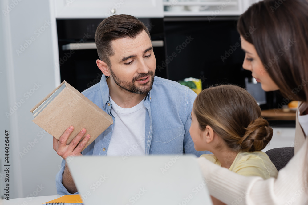 Obraz premium man holding textbook while helping daughter doing homework near wife and blurred laptop