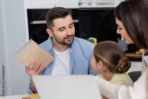man holding textbook while helping daughter doing homework near wife and blurred laptop