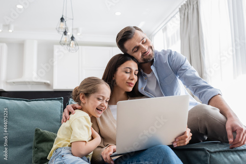 laughing girl watching movie on laptop near joyful parents smiling with closed eyes