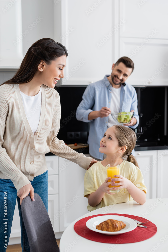 smiling girl with glass of orange juice looking at mom near blurred father mixing vegetable salad smiling girl with glass of orange juice looking at mom near blurred father mixing vegetable salad