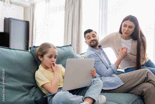 smiling parents looking at thoughtful daughter using laptop on sofa at home