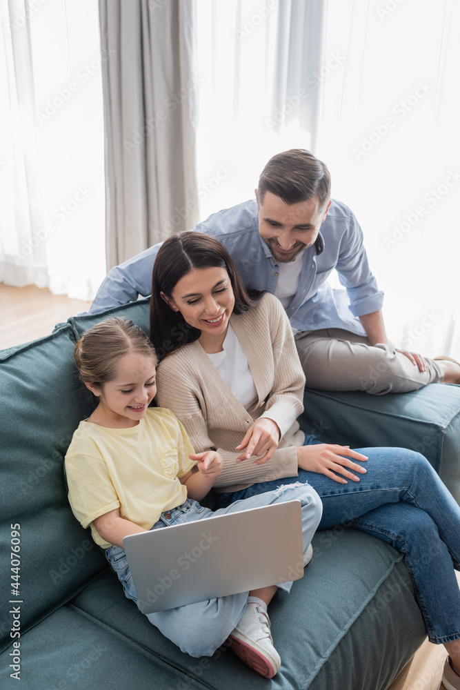 happy man near cheerful wife and daughter pointing at laptop while sitting on sofa happy man near cheerful wife and daughter pointing at laptop while sitting on sofa