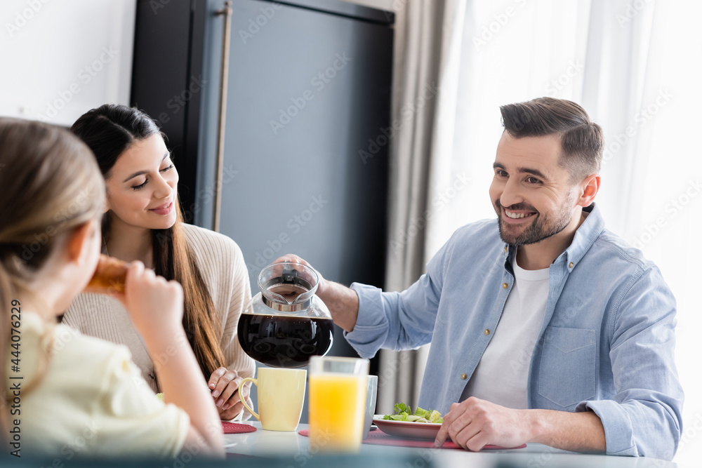 smiling man pouring coffee near wife and blurred daughter during breakfast smiling man pouring coffee near wife and blurred daughter during breakfast