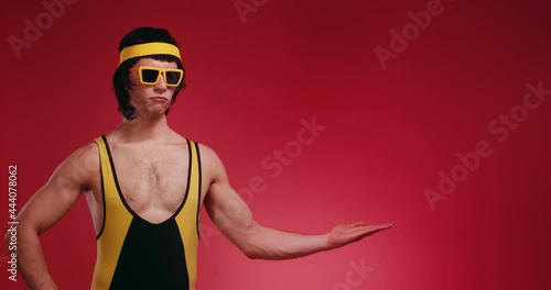 Funny retro hipster athlete man in yellow tracksuit and sunglasses from 80's show and represent product logo by his hands on a copy space isolated on red background. Promotion commercial concept