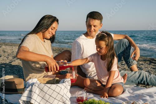 Family spending time together seaside outdoor. Picnic lunch with fruits. Happy people eating healthy food, drinking juice and sitting on blanket on the beach. Summer leisure concept.