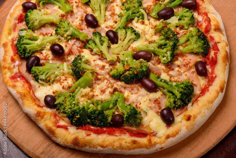 Broccoli pizza with black olives on wooden board and vegetables in the background.