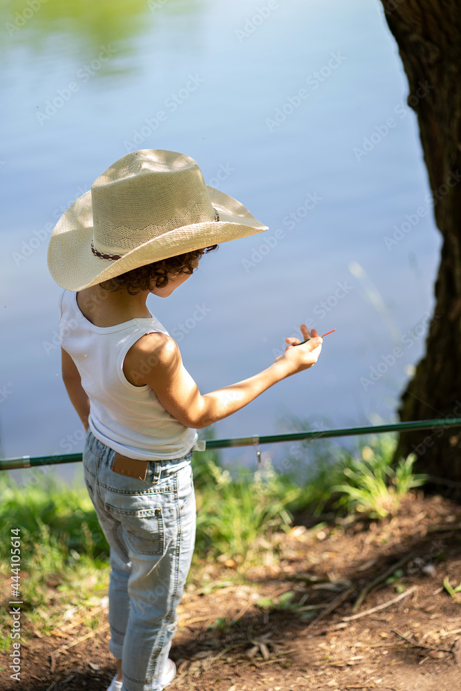 A boy in a straw hat holds a bobber by a fishing rod. Child fishing on a summer day.