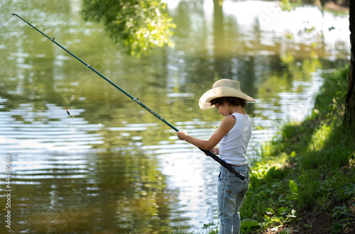 A boy with curly hair in a straw hat stands with a fishing rod by the pond. Child fishing on a summer day.