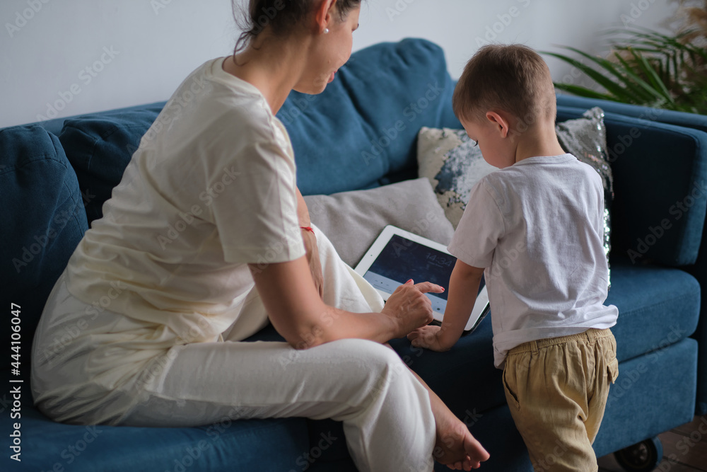 mother and son with tablet at home. Mother showing media content on line to her son in a tablet sitting on a couch in the living room in a house interior mother and son with tablet at home. Mother showing media content on line to her son in a tablet sitting on a couch in the living room in a house interior