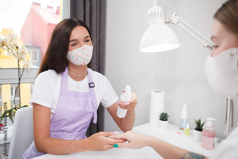 Professional manicurist wearing face mask, disinfecting hands of female client