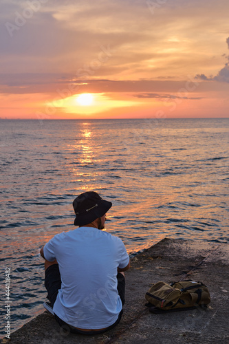 A young man sits by the sea and looks at the dawn. Male on travels. Amazing picturesque outdoor view. Beautiful dawn on the seashore. Healthy Lifestyle  Leisure Activities. Travel and Exploration