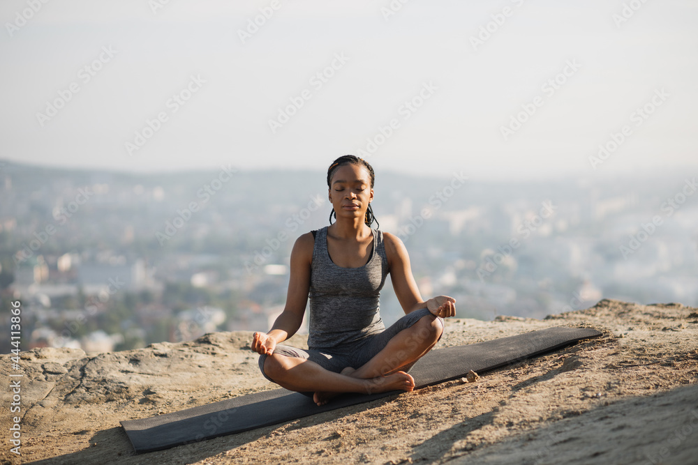 African american woman with closed eyes meditating in lotus position on yoga mat. Fitness young lady in sport clothes enjoying workout during morning time outdoors.