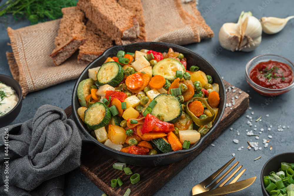 Vegetable stew in a frying pan on a gray background with bread and garlic.