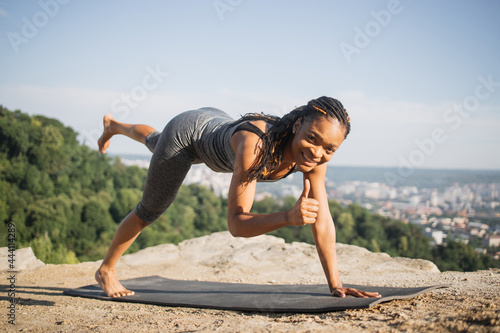 Cheerful african woman in sport clothes doing morning exercise on yoga mat outdoors, standing in Donkey, Kick fire hydrant exercise and showing thumb up. Healthy young lady training on fresh air.