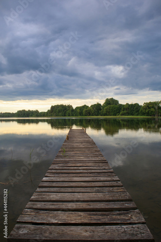 A long wooden footbridge enters the lake. Thick, dark rain clouds above the lake, sunset in the distance