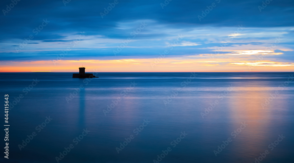 Image of Rocco Tower at St Ouens Bay, Jersey CI at sunset with silky water dark clouds with reflection of sunshine.