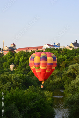 A large multi-colored balloon flies very low over the river and between dense trees. In the background, the buildings of the ancient city