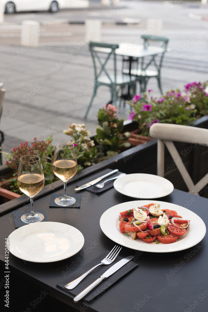 Italian caprese salad with chopped tomatoes, mozzarella, basil, oregano, thassos, onions, radishes, chili peppers and olive oil on a white plate. two glasses of wine rose in a restaurant on the street