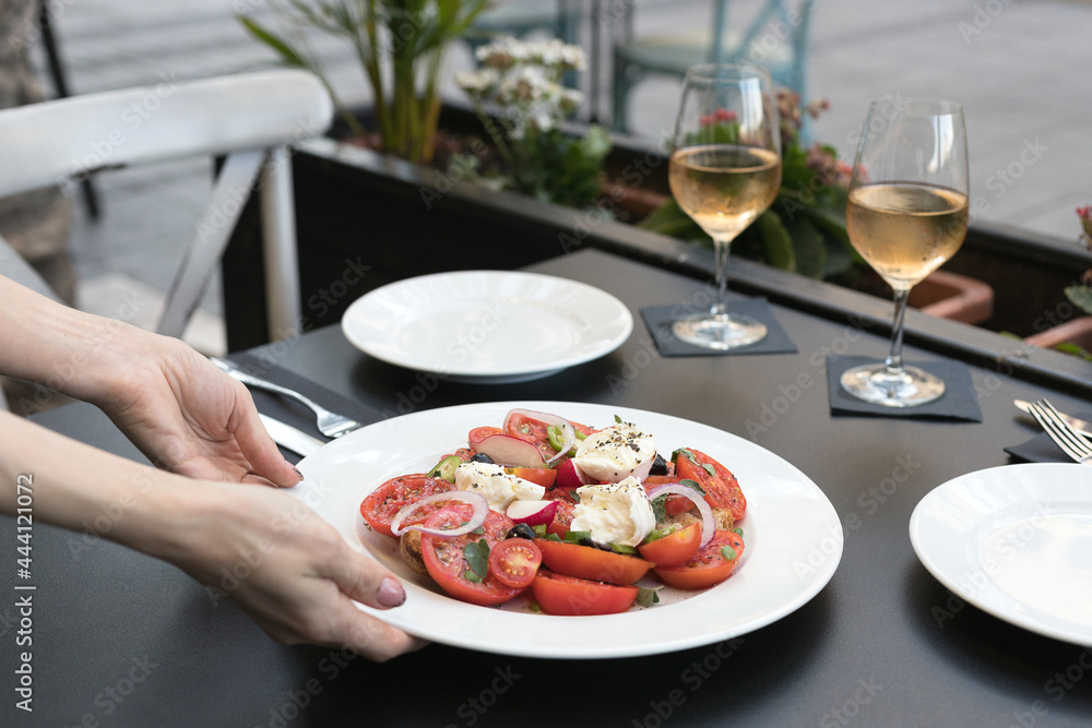 Italian caprese salad with tomatoes, mozzarella, oregano, thassos, onions, chili peppers and olive oil. two glasses of wine rose in a restaurant on the street. The waitress serves a dish