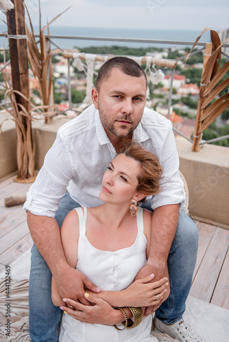 Close up portrait of the happy middle-aged couple in white hugs on the floor on open terrace of the roof overlooking the sea. Family is celebrating wedding anniversary. Interior In maracans, eco style