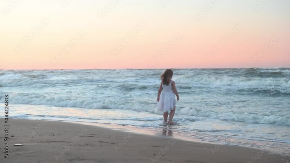 little girl child in a long sundress with long hair runs along the beach along the sea at sunset in summer. High quality 4k footage little girl child in a long sundress with long hair runs along the beach along the sea at sunset in summer. High quality 4k footage
