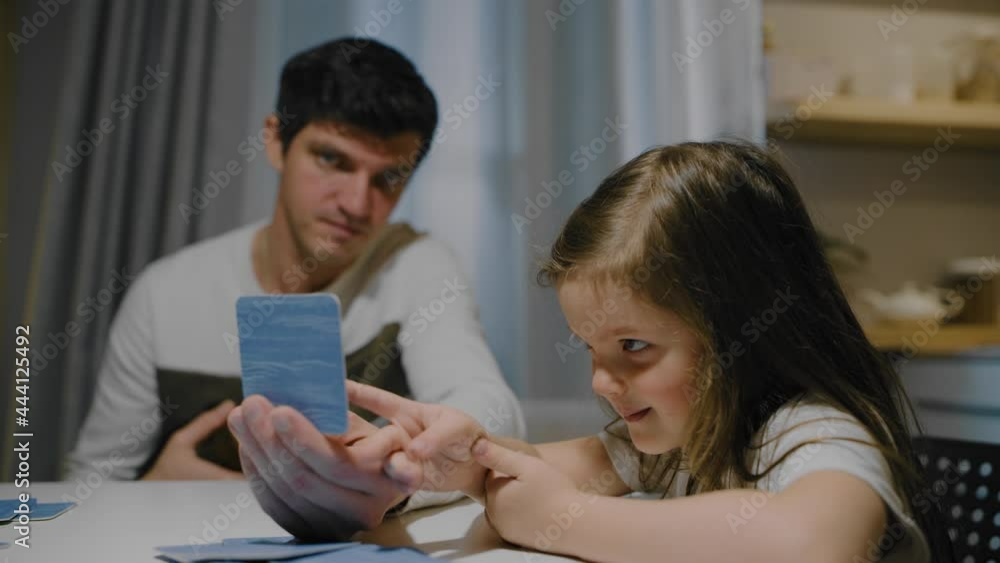 Father and daughter in the evening in the kitchen play board card games. The cat is sitting in Dad's arms. Father and daughter in the evening in the kitchen play board card games. The cat is sitting in Dad's arms.