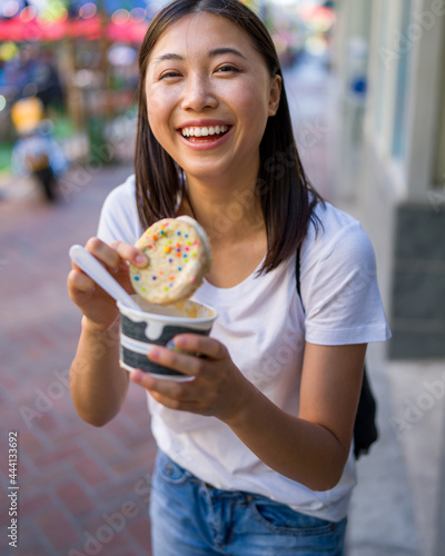 Happy Young Asian Woman Wearing Distressed Jeans and T-Shirt Eating an Ice Cream Sandwich in a Downtown Urban Area