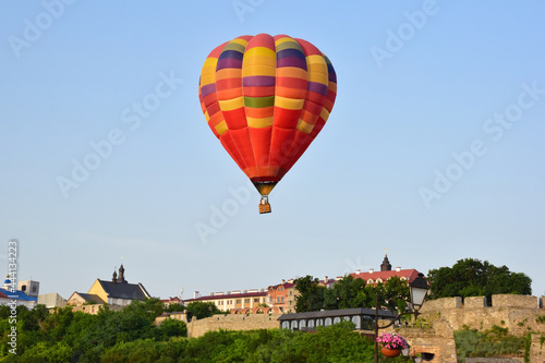 A large multicolored hot air balloon flies low over ancient buildings and a tree-lined valley. Photographed at close range