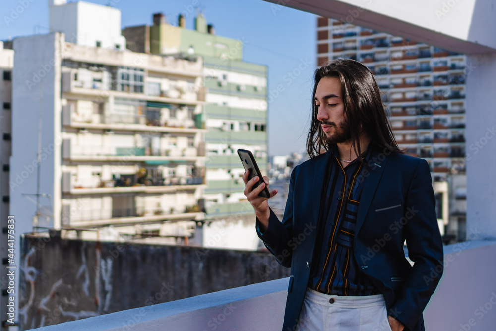 young Hispanic Latino businessman with long hair, using his smart phone on a terrace overlooking the city.