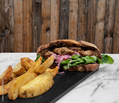 Fast food. Sandwich. Closeup view of a pork meat bondiola sandwich with lettuce, onion pickles and fried potatoes in a black dish on the pub white marble table.  photo