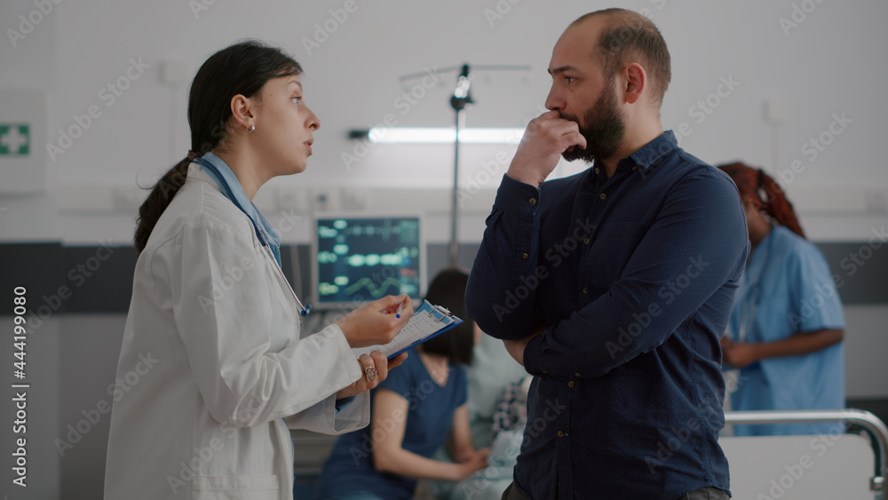 Fototapeta premium Pediatric woman doctor explaining disease symptoms to father while black nurse writing medical sickness treatment on clipboard in hospital ward. Sick little child resting in bed during examination