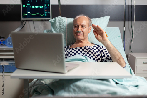 Portrait of senior man greeting family waving at laptop camera laying in hospital bed, after illness diagnosis, breathing with oxygen tube. Modern eqipment monitoring pacient heart rate during photo