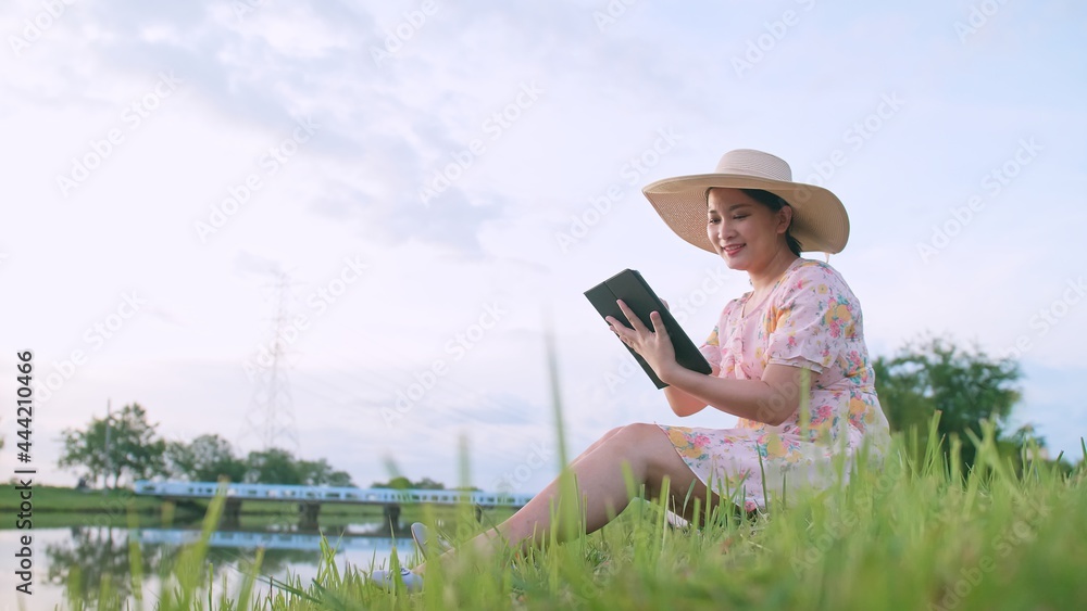 Asian woman relaxing with using portable device sitting on the lawn near the river.