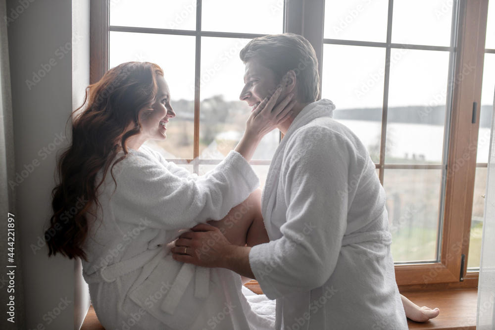 A couple in white bathrobes standing near the window and looking romantic A couple in white bathrobes standing near the window and looking romantic