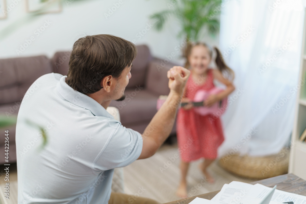 father works at home with laptop together with his daughter. little girl wants to play guitar, noise and interfere with work. man shakes his fist and asks them to calm down with remote work at home father works at home with laptop together with his daughter. little girl wants to play guitar, noise and interfere with work. man shakes his fist and asks them to calm down with remote work at home