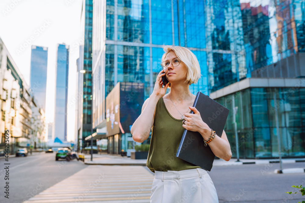 Young business woman standing with laptop among the skyscrapers and speaks on the phone.