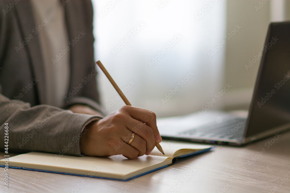 Female hand writes in the notepad near the laptop. Married woman in jacket working at home office. Horizontal view. Female hand writes in the notepad near the laptop. Married woman in jacket working at home office. Horizontal view.