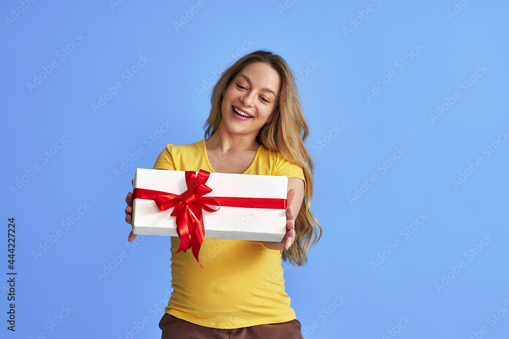 attractive pregnant woman in a yellow T-shirt pulls forward a gift box with a red flight card standing on a blue background. Preparing for holiday, giving a gift. Advertising discounts and promotions attractive pregnant woman in a yellow T-shirt pulls forward a gift box with a red flight card standing on a blue background. Preparing for holiday, giving a gift. Advertising discounts and promotions