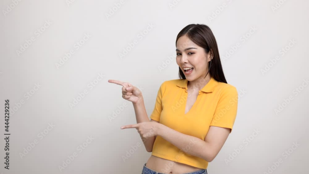 Excited asian woman pointing the finger to blank space for advertising text on isolated background. Joyful teenage girl in yellow shirt standing in white room looking at empty space. Excited asian woman pointing the finger to blank space for advertising text on isolated background. Joyful teenage girl in yellow shirt standing in white room looking at empty space.