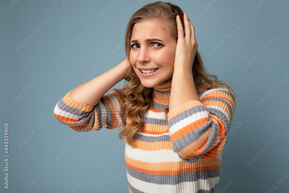 Photo of young emotional angry pretty nice blonde curly woman with sincere emotions wearing casual striped pullover isolated on blue background with free space and covering ears trying not to hear Photo of young emotional angry pretty nice blonde curly woman with sincere emotions wearing casual striped pullover isolated on blue background with free space and covering ears trying not to hear