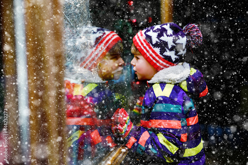 Cute little healthy school kid boy on Christmas market. Funny happy child in fashion winter clothes making window shopping decorated with gifts, xmas tree. Snow falling down, snowfall