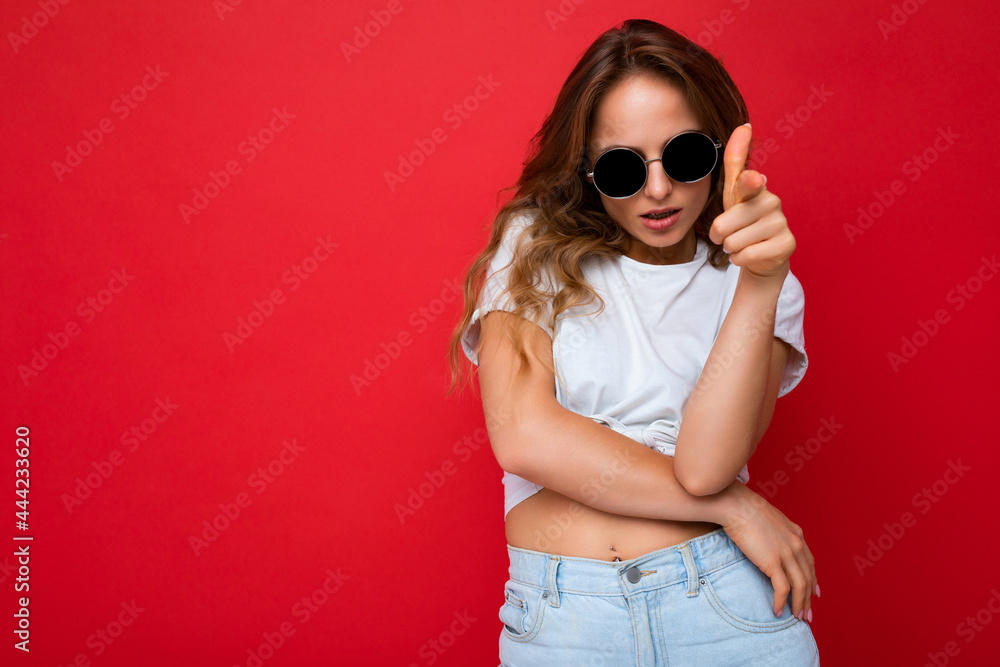 Shot of beautiful self-confident young dark blonde curly woman isolated over red background wall wearing casual white t-shirt and stylish sunglasses looking at camera and pointing finger at you