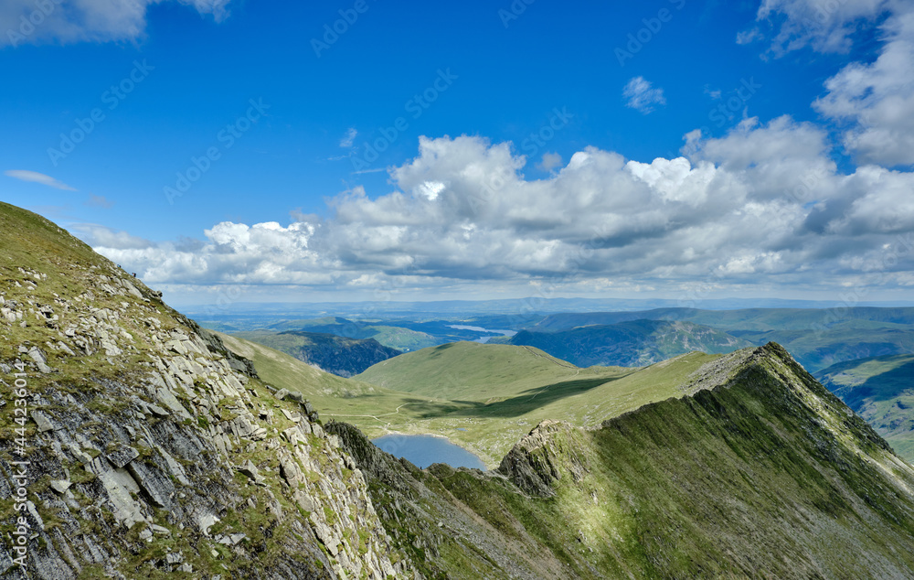 Red Tarn peeking over Striding Edge, on Helvellyn in the Lake District shot on a summer day with blue skies and white clouds, with Ullswater in the far distance