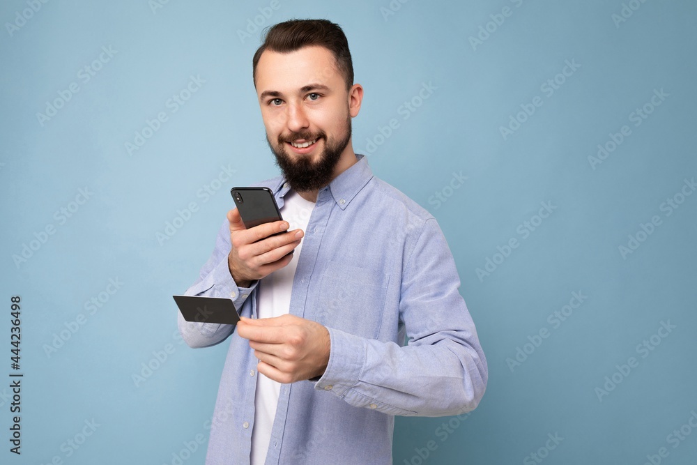 Handsome man wearing everyday clothes isolated on background wall holding and using phone and credit card making payment looking at camera Handsome man wearing everyday clothes isolated on background wall holding and using phone and credit card making payment looking at camera