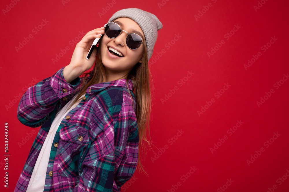 photo of beautiful happy positive young blonde woman wearing hipster purple shirt and casual white t-shirt grey hat and sunglasses isolated over red background holding in hand and speaking on mobile