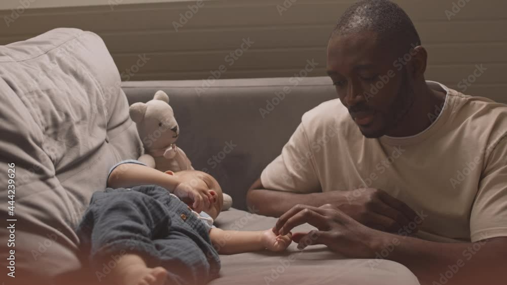 Medium close-up of caring African man sitting by couch at home, touching tiny hand of peaceful Mixed-Race baby who sleeping on sofa Medium close-up of caring African man sitting by couch at home, touching tiny hand of peaceful Mixed-Race baby who sleeping on sofa