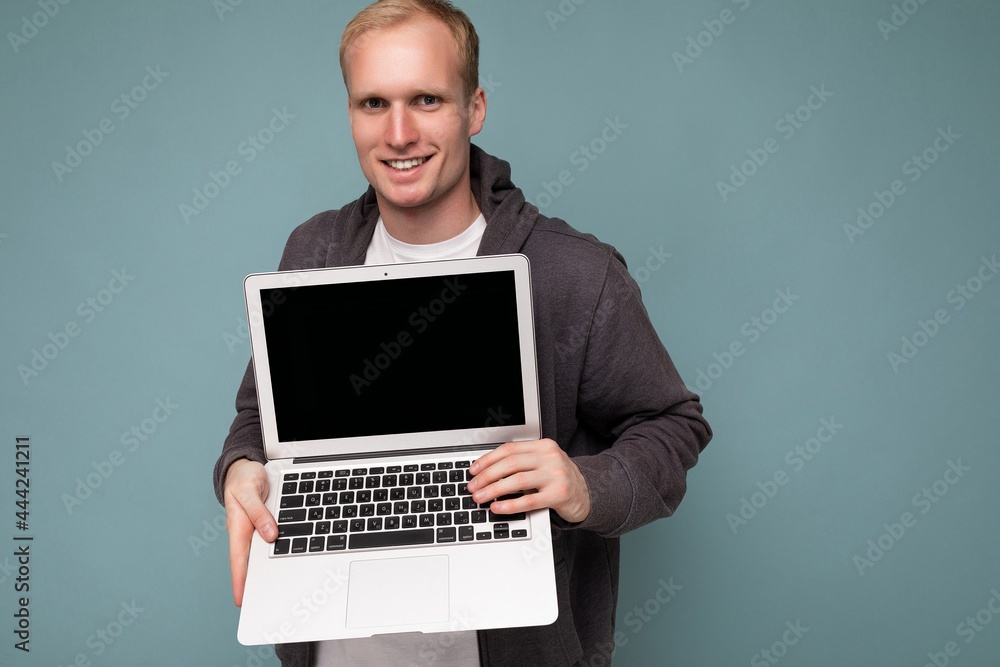 Close-up portrait of handsome smiling blonde man holding computer laptopwith empty monitor screen with mock up and copy space looking at camera isolated over blue background Close-up portrait of handsome smiling blonde man holding computer laptopwith empty monitor screen with mock up and copy space looking at camera isolated over blue background