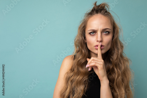 Photo of young beautiful blonde curly woman with sincere emotions wearing black top isolated over blue background with copy space and showing silence gesture with finger over lips