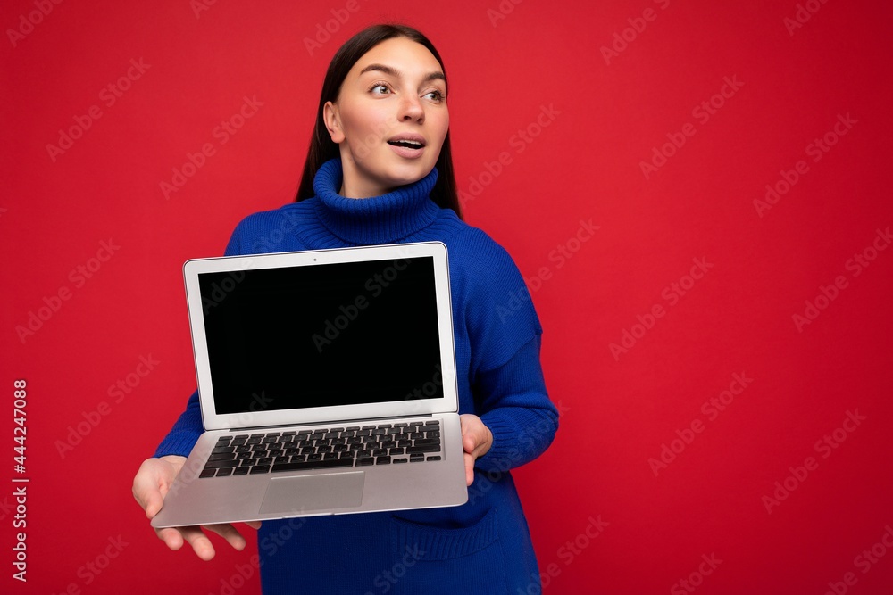 Fascinating astonished pretty happy beautiful young brunette female person holding computer laptop looking to the side wearing dark blue sweater isolated over red wall background Fascinating astonished pretty happy beautiful young brunette female person holding computer laptop looking to the side wearing dark blue sweater isolated over red wall background